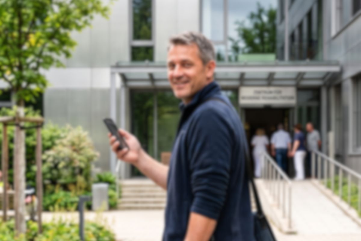 Mann mit Gehstock und Smartphone vor dem Klinikgebäude